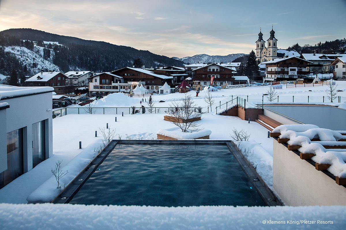 Ein dampfender Infinity-Pool im Vordergrund einer tief verschneiten alpinen Hotellandschaft. Im Hintergrund erstreckt sich ein malerisches Dorf mit traditionellen Holzhäusern, einer Doppelturmkirche und schneebedeckten Bergen in der Dämmerung.
