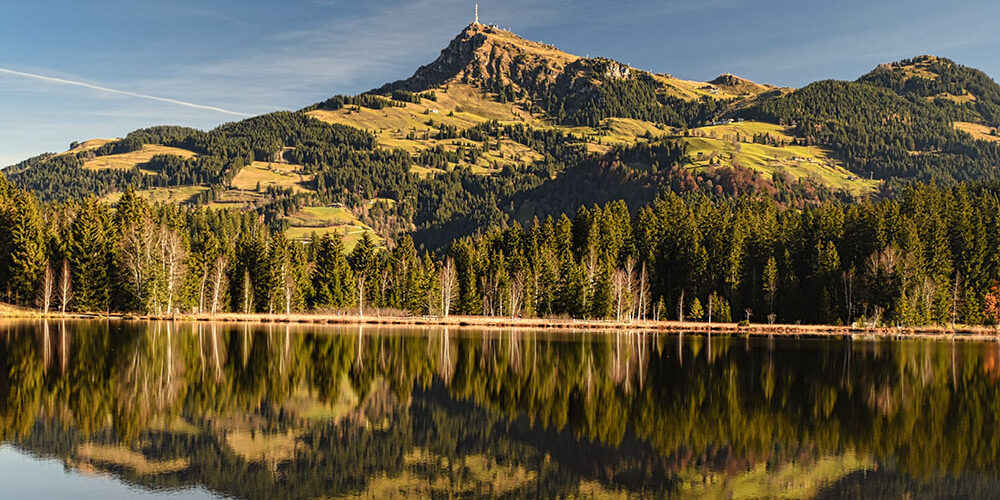 Landschaftsaufnahme der Kitzbüheler Alpen im warmen Sonnenlicht: Dichte grüne Wälder und das Kitzbüheler Horn spiegeln sich spiegelglatt auf der Oberfläche eines idyllischen Bergsees unter blauem Himmel.