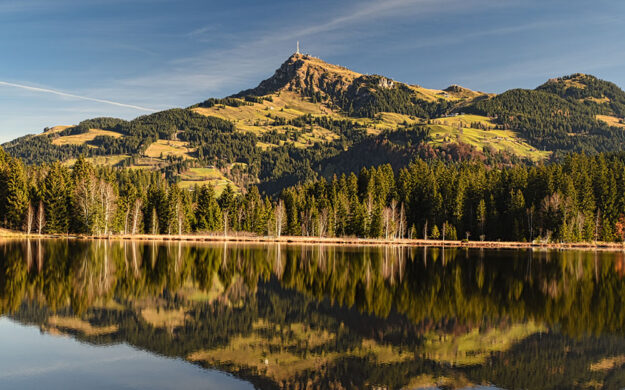 Kitzbüheler Alpen Blog Landschaftsaufnahme der Kitzbüheler Alpen im warmen Sonnenlicht: Dichte grüne Wälder und das Kitzbüheler Horn spiegeln sich spiegelglatt auf der Oberfläche eines idyllischen Bergsees unter blauem Himmel.