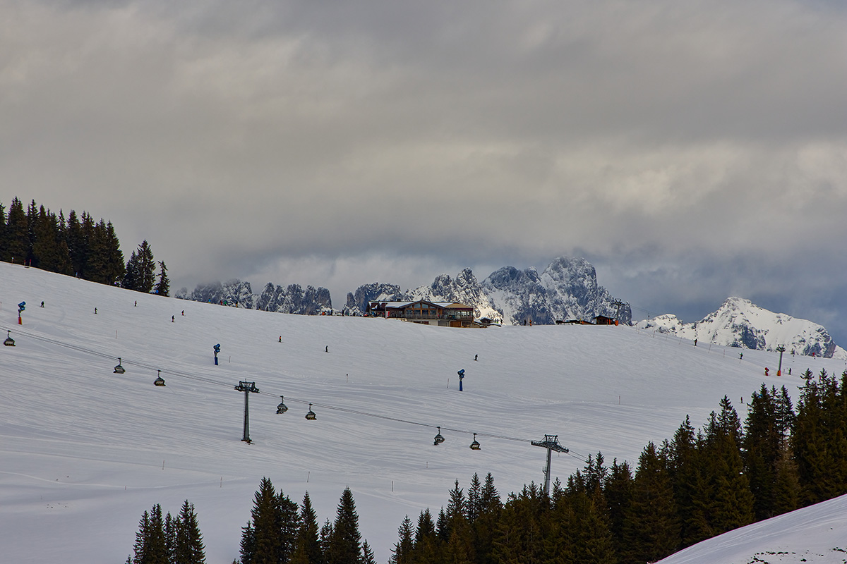 WilderKaiserBlog_4 Winterlandschaft in den Sextner Dolomiten, Südtirol, mit Skigebiet Drei Zinnen Dolomiten. Blick auf eine weite Skipiste mit Sessellift-Anlagen, auf der Skifahrer unterwegs sind. Auf einem Hügel steht ein Bergrestaurant-Gebäude mit Holzfassade. Im Hintergrund ragen markante, verschneite Felsgipfel unter einem dramatischen, bewölkten Himmel empor.