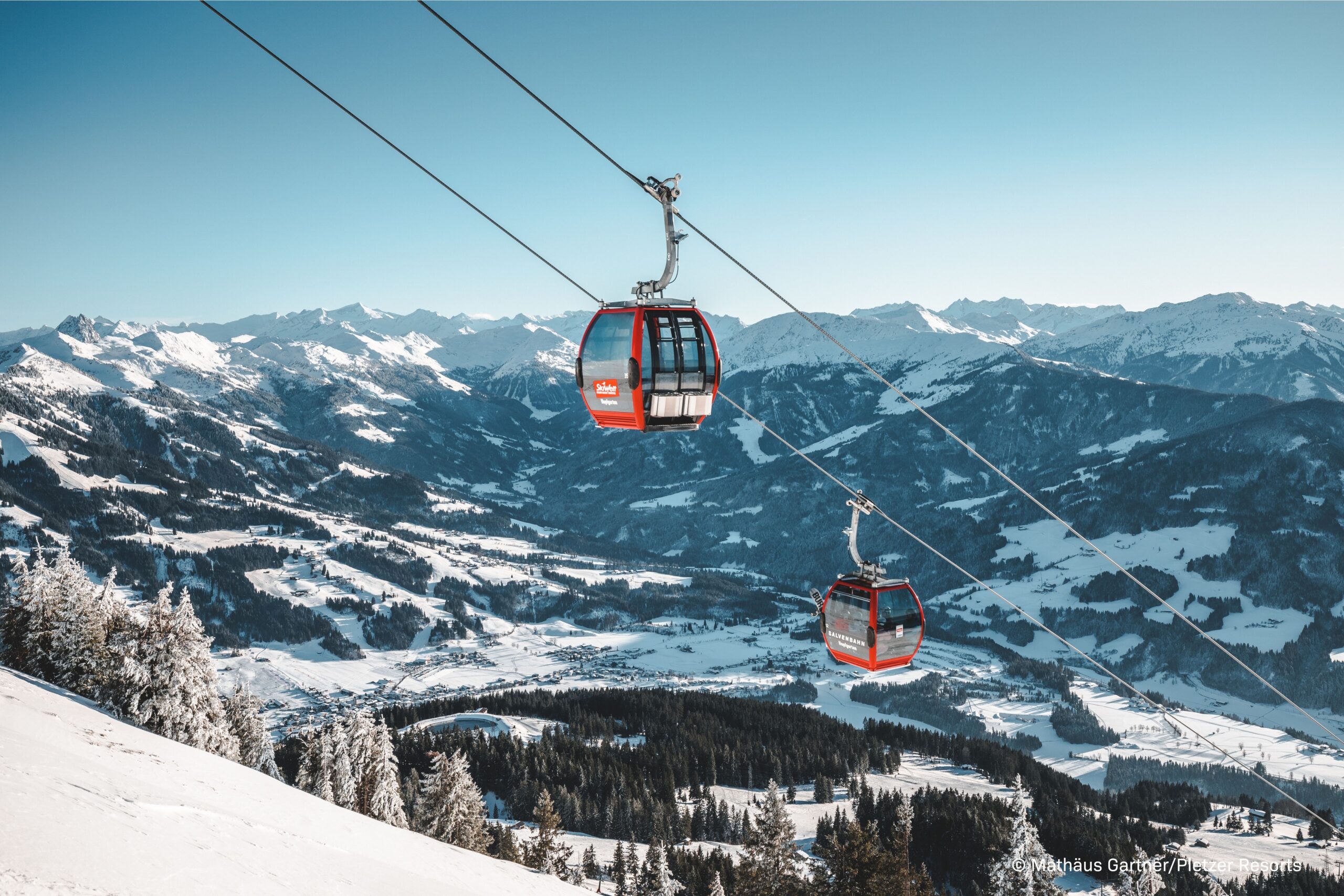 Panoramaaufnahme im Wintersportgebiet: Eine moderne rot-schwarze Gondelbahn schwebt bei klarem, blauem Himmel hoch über einem verschneiten Tal und tief verschneiten Tannenwäldern.